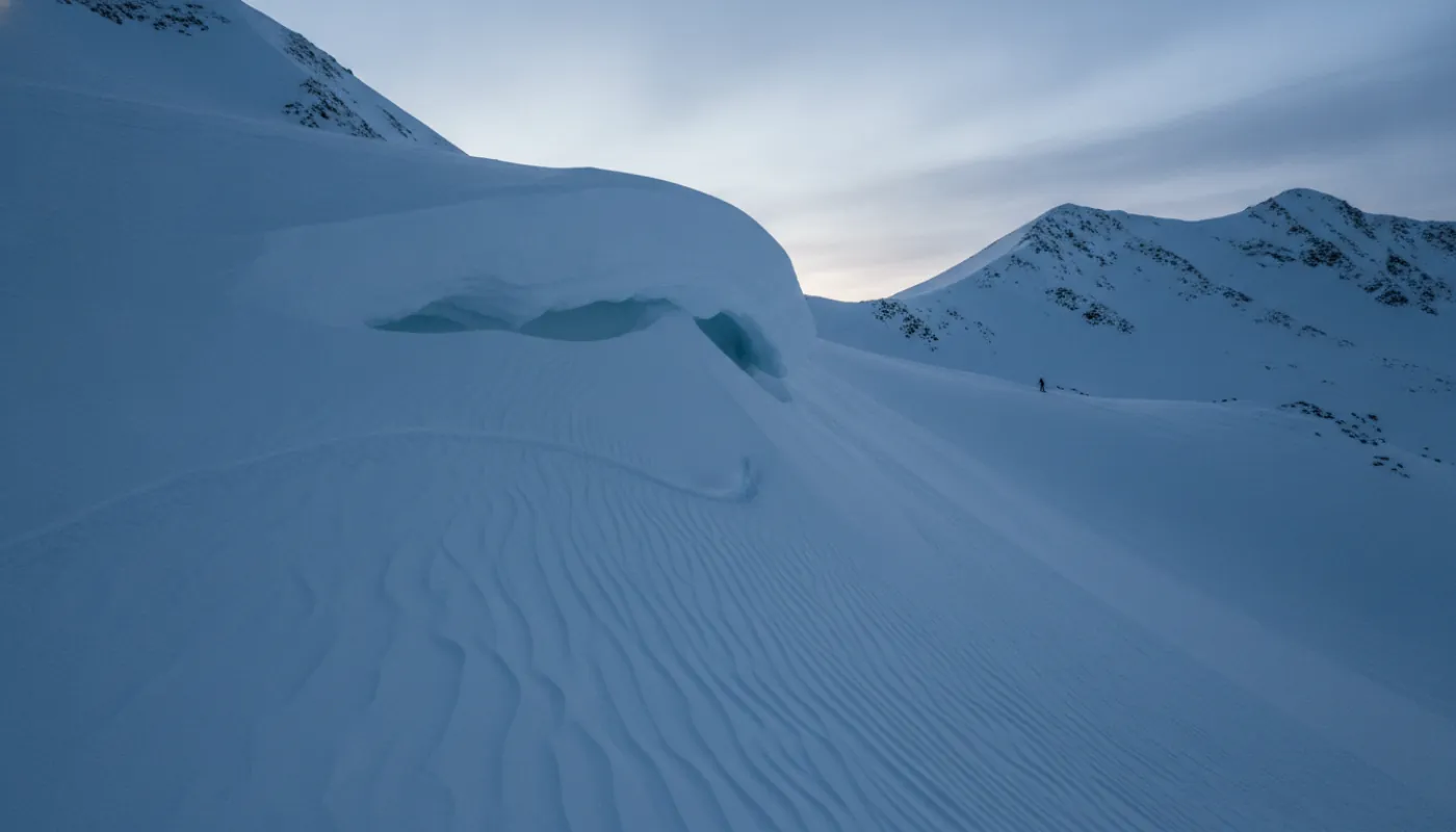 A wind-loaded backcountry slope at first light, a cornice curling over a steep north-facing bowl streaked with wind texture, a small recent fracture line visible in the snow, cold flat blue light