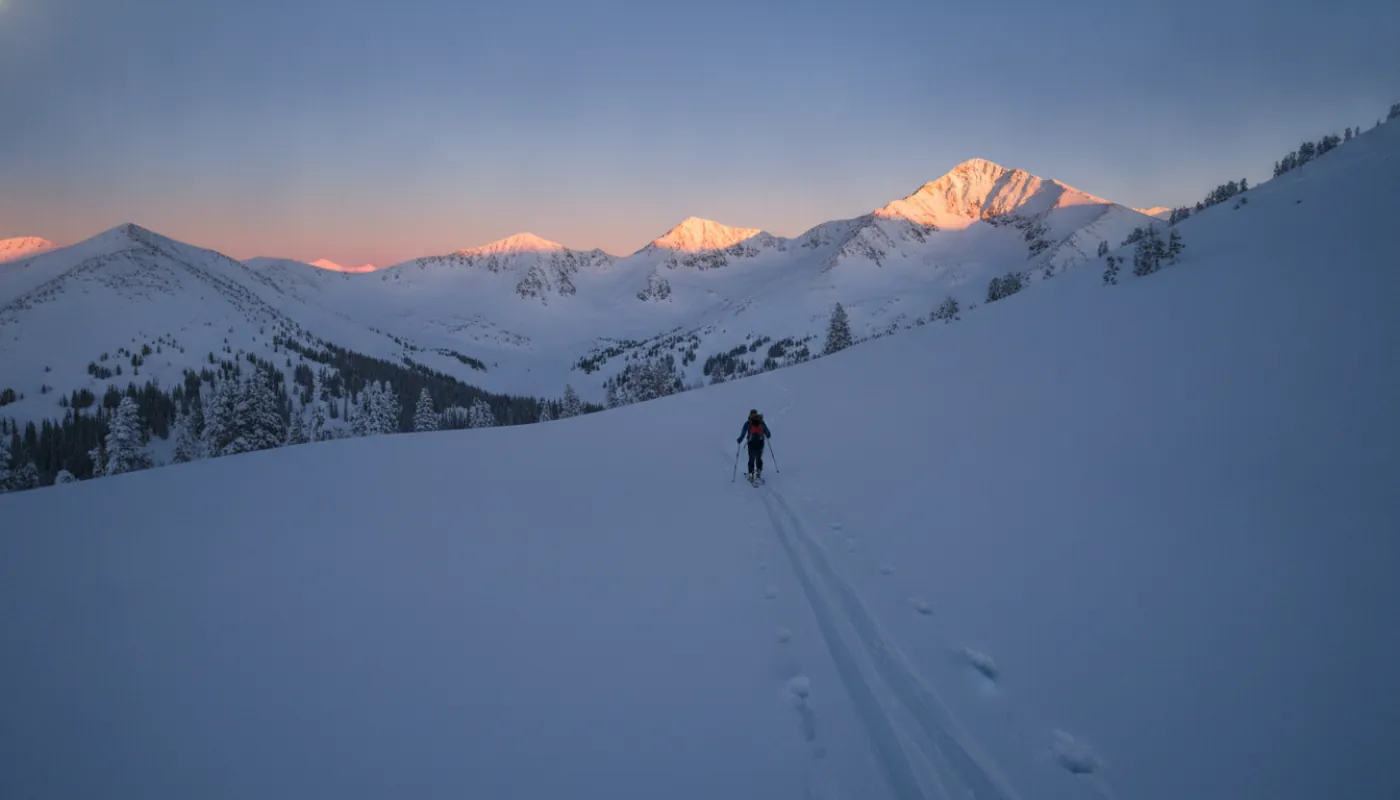 A backcountry skier climbing a skin track through open snow at dawn, skins on the ski bases, herringbone tracks trailing downhill behind, long cold shadows across the slope as the peaks catch first light